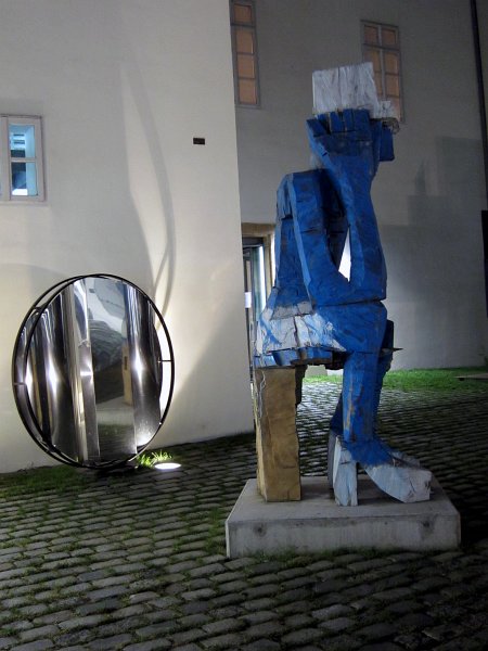 IMG_2880_1.jpg - Georg Baselitz - Volk Ding Zero. Skulptura Volk Ding Zero – Folk Thing Zero (2009) Georga Baselitze (nar. 1938) je tři metry vysoký bronz, který byl odlit na jaře roku 2009 podle Baselitzovy vlastní pomalované skulptury z cedrového dřeva. Asi půldruhé tuny těžká patinovaná bronzová socha představuje muže pomalovaného modrou a bílou olejovou barvou, který sedí na stoličce s nohama v klasických dámských lodičkách s vysokými podpatky. Na hlavě má tento muž, zamyšleně se dotýkající pravou ruku spánku, bílou čepici, která svým tvarem připomíná papírovou čepici, jakou si člověk sám zhotovuje z novin jako ochranu proti kapkám barvy. Skulptura má syrový, expresivní výraz připomínající hrubou kresbu. Její hlava je vlastní podobiznou umělce, první tohoto druhu v bronzu.