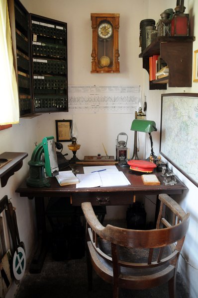 DSC_7582_1.jpg - Dopravní kancelář z poválečného období. Na stole nalezneme: Dopravní deník, telefon, razítka, kalamář s pérem, kompostér, elektrická lampa. Na levé zdi visí: ternión na jízdenky, dole jsou zavěšeny: ruční návěstidla, plombovací kleště. Vpravo visí mapa ČSD z roku 1947 a nad stolem jsou zavěšeny kyvadlové hodiny.